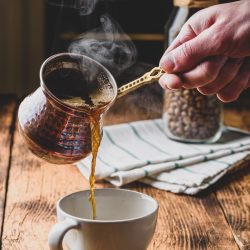 Pouring turkish coffee into white cup. Jar of roasted coffee beans on background
